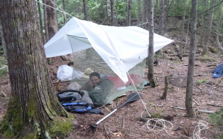 a person smiles from under a tarp shelter amongst trees on an outward bound course